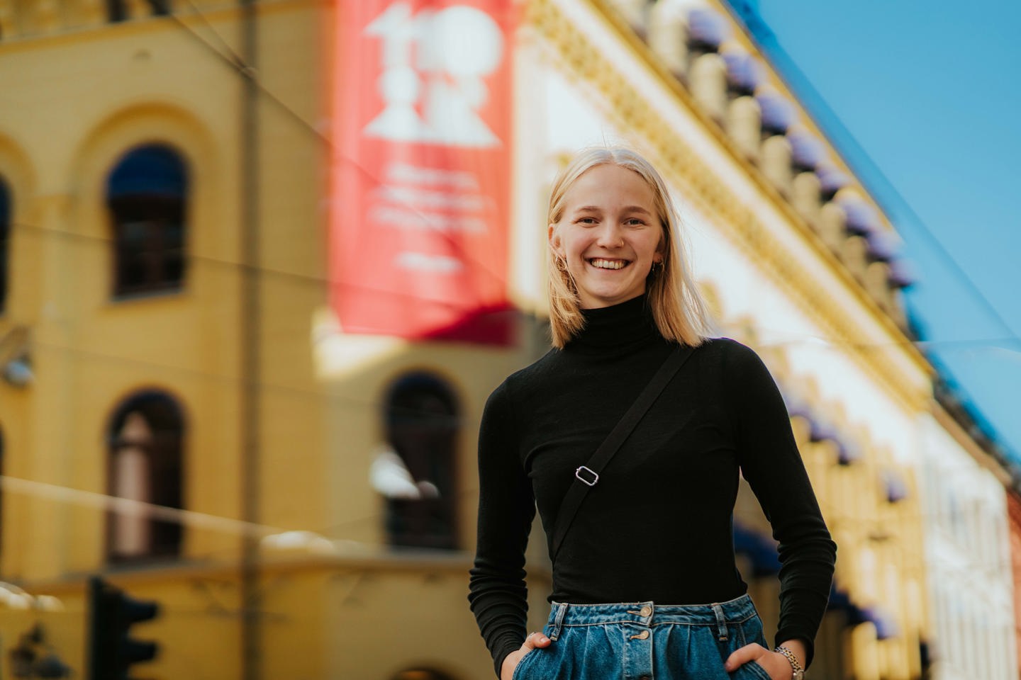 En smilende student står foran campus. Hun har blondt hår og bærer en sort høyhalset t-skjorte