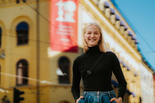 En smilende student står foran campus. Hun har blondt hår og bærer en sort høyhalset t-skjorte