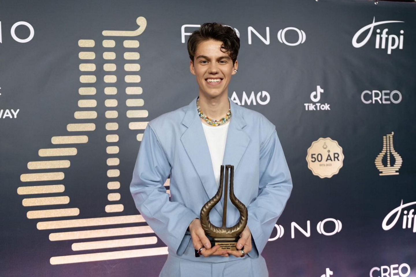 Peder Elias står foran en fotovegg på Spellemannsprisutdelingen. Han holder Spellemannstroféet og smiler bredt. Han har på seg lyseblå dress, hvit t-skjorte og fargerike halssmykker av krystaller og andre mineraler..