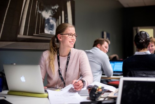 Kvinne med briller og rosa genser som sitter foran en laptop