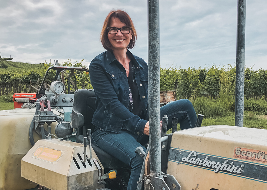 Solfrid Lind sitter oppå en gul traktor. Hun har på seg en olajakke og olabukser, sorte briller og har kort brunt hår.