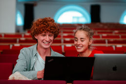 To smilende studenter, en mann og en kvinne, sitter med hver sin laptop i et auditorium. De ser ut til å ha det gøy mens de studerer.