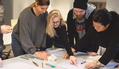 Foto av fire studenter ved et bord som peker og tegner på et ark med skisser.