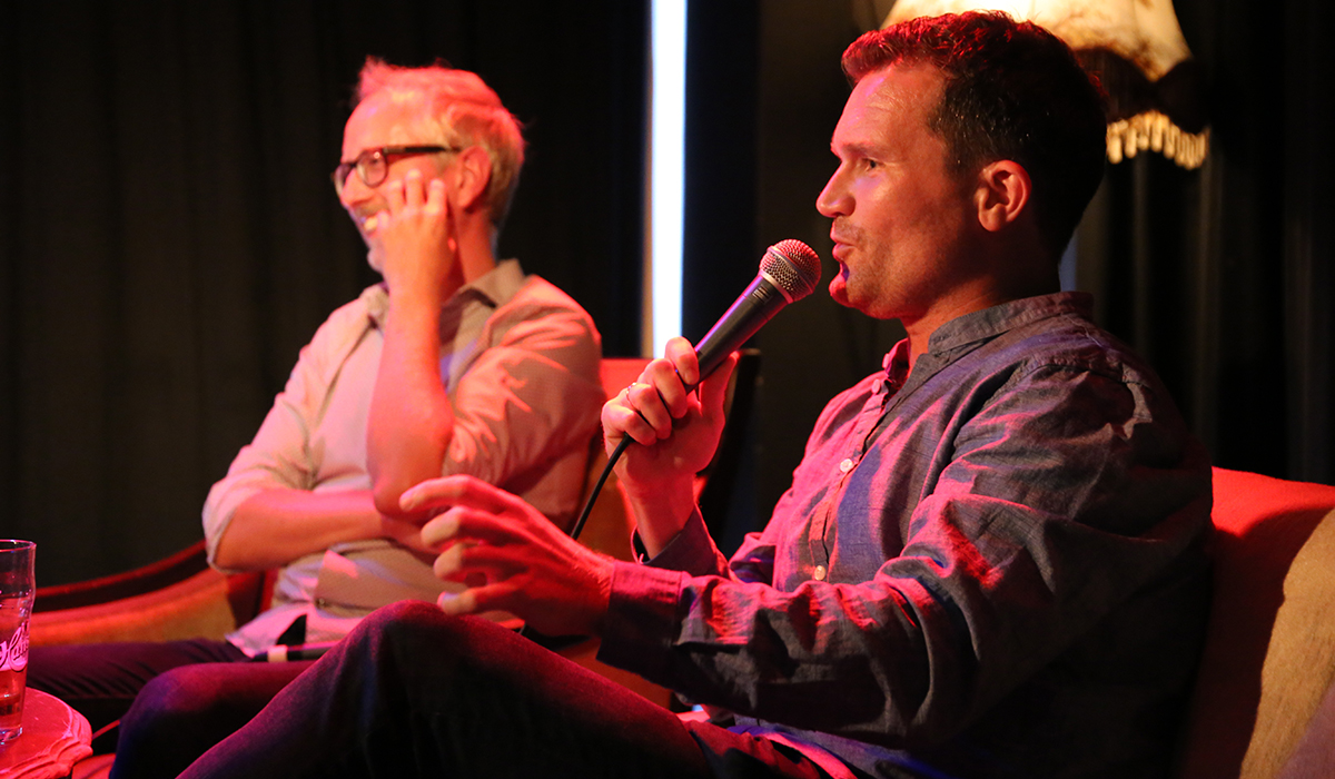 Bård Vegar Solhjell og Ketil Raknes sitter engasjert i samtale med et rødt scenelys. Bak dem er et sort sceneteppe. De sitter i retro stoler. 
