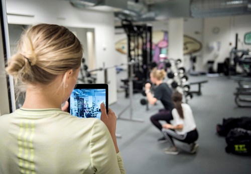 Studenter som trener i treningsstudio, hvor en av dem filmer med ipad