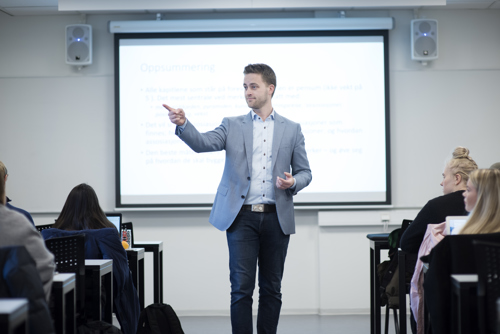 Foreleser som står vendt mot klassen, med en skjerm bak seg,