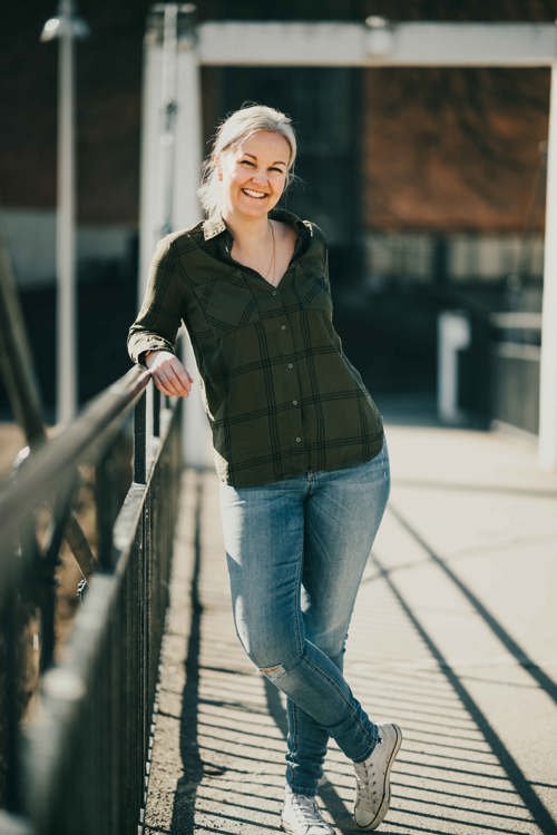 Voksen, blond dame i grønn skjorte og blå jeans lener seg på rekkverk.