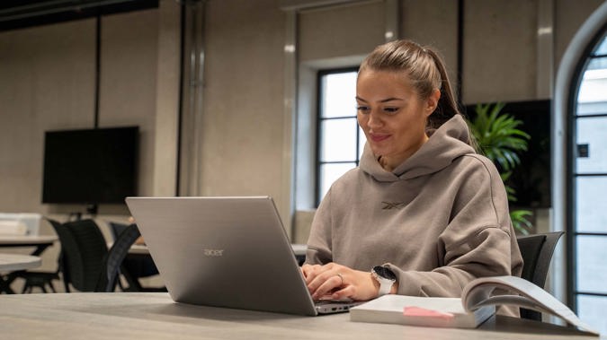 En student sitter ved et bord og skriver på en pc. Ved siden av pc-en ligger det en åpen bok.